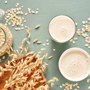 Oat milk in a glass and mug on a blue background. Flakes and ears for oatmeal and granola on a wooden plate. Vegan ingredient solutions for dairy replacement.